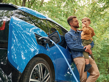 Das Bild zeigt ein E-Auto, davor ein Vater, der sein Kind in den Armen hält.